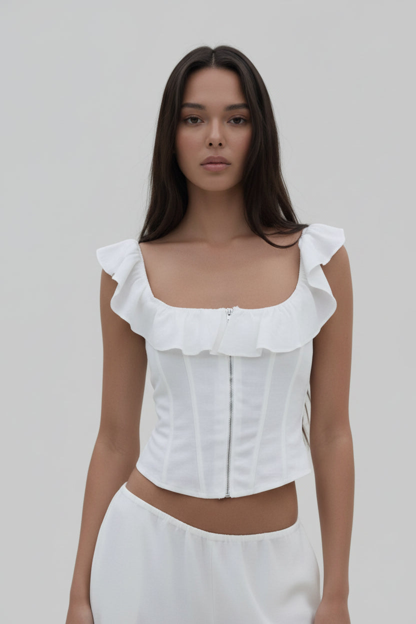 Person wearing a white ruffled top and pants in an outdoor setting with greenery.