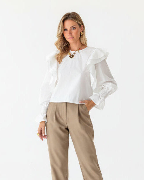 Woman wearing a white blouse with ruffled sleeves and beige pants against a wooden background