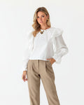 Woman wearing a white blouse with ruffled sleeves and beige pants against a wooden background